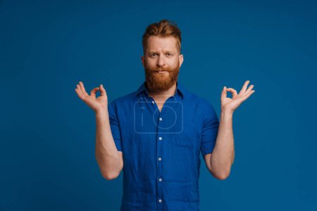 Téléchargez les photos : Portrait d'adulte beau rousse élégant barbu homme calme en chemise bleue faisant geste de méditation et regardant la caméra, tout en se tenant sur fond bleu isolé - en image libre de droit