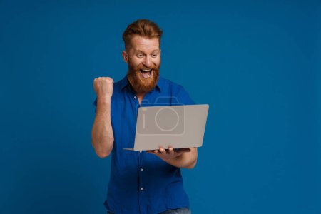 Téléchargez les photos : Adulte beau rousse barbu homme enthousiaste avec la bouche ouverte tenant ordinateur portable et faisant geste gagnant, tout en se tenant sur fond bleu isolé - en image libre de droit