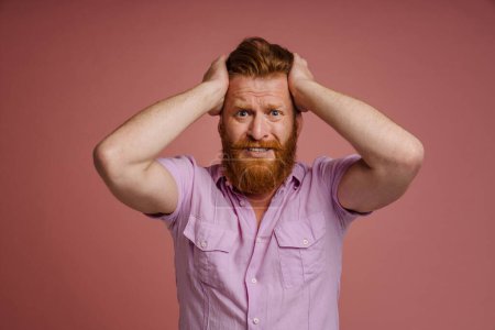 Téléchargez les photos : Adult handsome stressed redhead bearded man in pink shirt holding head and looking at camera, while standing over isolated coral background - en image libre de droit