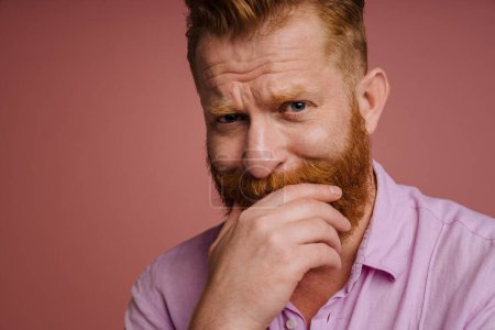 Foto de Cropped portrait of adult sad redhead bearded man covering his mouth and looking at camera , while standing over isolated coral background - Imagen libre de derechos