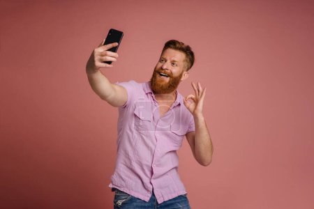 Photo for Adult handsome stylish redhead bearded man taking selfie on phone and doing OK gesture, while standing over isolated coral background - Royalty Free Image