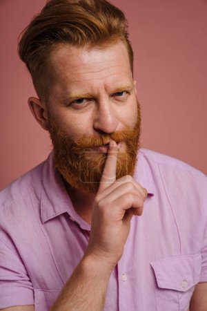 Téléchargez les photos : Adult handsome redhead bearded frowning man doing silence gesture with finger on lips and looking at camera, while standing over isolated coral background - en image libre de droit