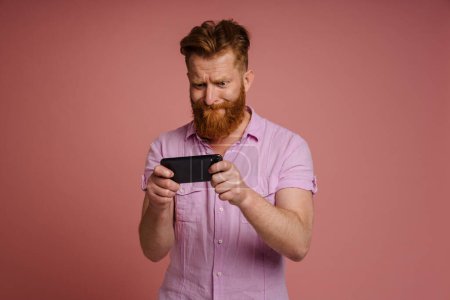 Téléchargez les photos : Adulte élégant rousse barbu concentré homme tendu en chemise rose jouer jeu mobile sur le téléphone, tout en se tenant debout sur fond de corail isolé - en image libre de droit