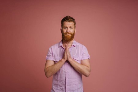 Téléchargez les photos : Adulte beau rousse barbu homme en chemise rose avec les mains pliées faire un geste de prière et en regardant la caméra, tout en se tenant sur fond de corail isolé - en image libre de droit