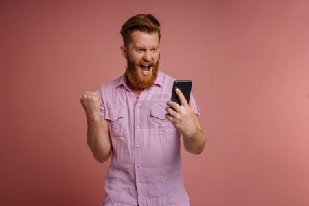 Photo for Adult handsome stylish redhead bearded enthusiastic man in pink shirt with raised fist and opened mouth holding phone and looking on it, while standing over isolated coral background - Royalty Free Image