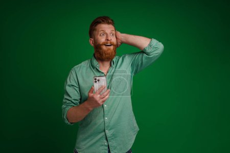 Téléchargez les photos : Ginger white man expressing surprise and using mobile phone isolated over green background - en image libre de droit