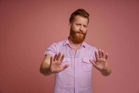 Foto de Jengibre disgustado hombre con barba mostrando gesto de parada en la cámara aislado sobre fondo rosa - Imagen libre de derechos
