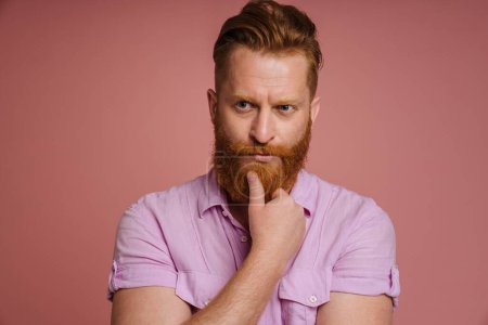 Photo for Ginger white man with beard looking aside and holding his chin isolated over pink background - Royalty Free Image