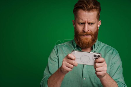 Photo for Ginger perplexed man in earphones playing online game on cellphone isolated over green background - Royalty Free Image
