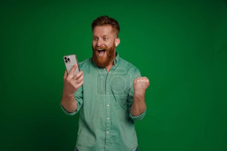 Photo for Ginger excited man in earphones playing online game on mobile phone isolated over green background - Royalty Free Image