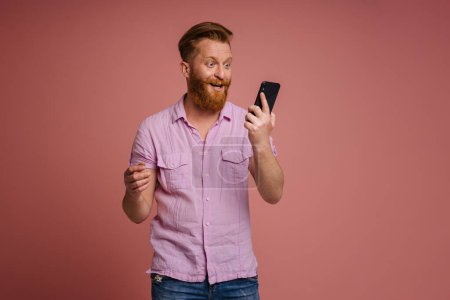 Téléchargez les photos : Ginger white man expressing surprise and using mobile phone isolated over pink background - en image libre de droit