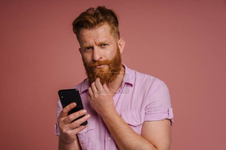 Photo for Ginger white man with beard frowning and using mobile phone isolated over pink background - Royalty Free Image