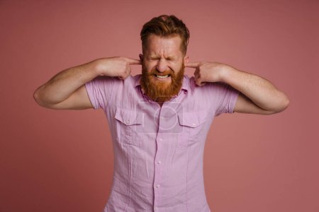 Photo for Ginger displeased man grimacing while plugging his ears isolated over pink background - Royalty Free Image