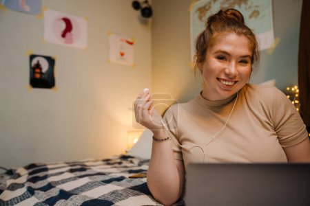 Foto de Retrato de una joven hermosa chica sonriente con portátil con auriculares en la mano y mirando a la cámara, mientras está sentada en una acogedora habitación en casa - Imagen libre de derechos