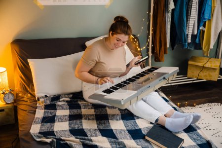Téléchargez les photos : Jeune belle fille souriante et heureuse jouant du piano électronique avec le téléphone à la main, tout en étant assise sur le lit dans une chambre confortable à la maison - en image libre de droit