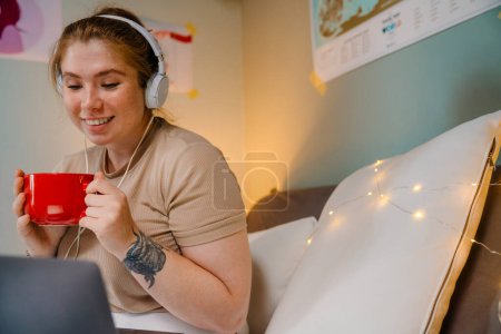 Foto de Joven hermosa chica sonriente en auriculares con taza roja mirando a la computadora portátil, mientras está sentado en una habitación acogedora en casa - Imagen libre de derechos