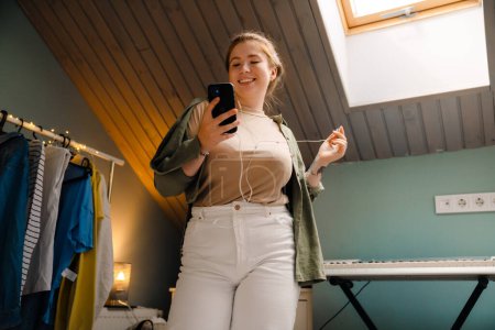 Photo for Young beautiful smiling happy girl in headphones with raised hand holding and using her phone , while standing in cozy room at home - Royalty Free Image