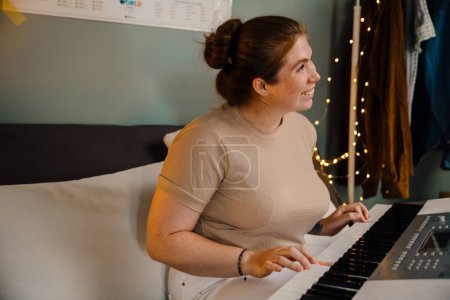 Téléchargez les photos : Jeune belle fille souriante heureuse jouant du piano électronique et regardant de côté, tout en étant assis à la maison - en image libre de droit
