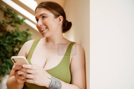 Téléchargez les photos : Gingembre jeune femme riant et utilisant un téléphone portable après la pratique du yoga à l'intérieur - en image libre de droit