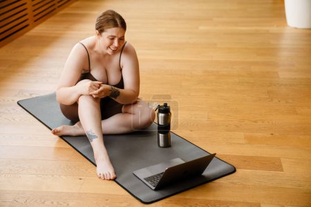 Téléchargez les photos : Gingembre jeune femme souriant et utilisant un ordinateur portable pendant la pratique du yoga à l'intérieur - en image libre de droit