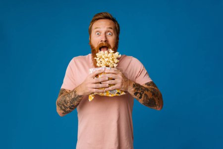 Photo for Shocked bearded white man in casual wear holding bucket with flying popcorn isolated over blue studio background - Royalty Free Image