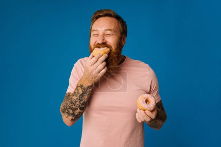 Téléchargez les photos : Homme barbu affamé en tenue décontractée manger des beignets isolés sur fond bleu studio - en image libre de droit