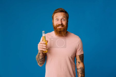 Foto de Retrato del hombre barbudo jengibre excitado sosteniendo cerveza mientras está de pie aislado sobre fondo de estudio azul - Imagen libre de derechos