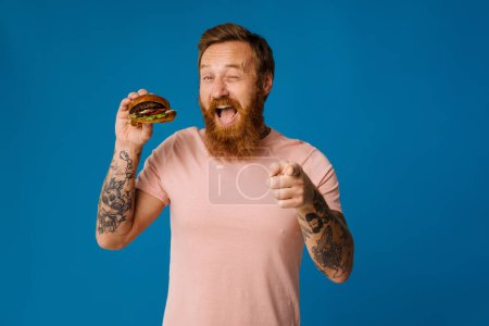 Téléchargez les photos : Blanc affamé homme barbu manger hamburger isolé sur fond bleu - en image libre de droit