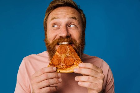 Foto de Barbudo feliz hombre comer pizza mientras de pie aislado sobre fondo azul - Imagen libre de derechos