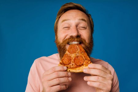 Téléchargez les photos : Barbu homme heureux manger de la pizza tout en se tenant isolé sur fond bleu - en image libre de droit
