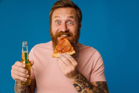 Photo for Bearded happy man holding pizza slice and beer while standing isolated over blue background - Royalty Free Image