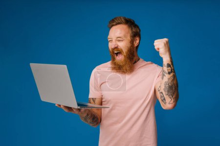 Foto de Barbudo hombre feliz gritando y sosteniendo el ordenador portátil, mientras que de pie aislado sobre fondo azul - Imagen libre de derechos