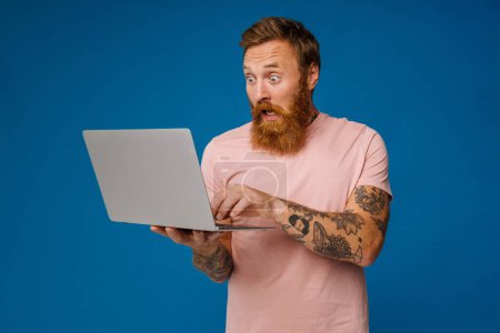 Téléchargez les photos : Barbu homme heureux tenant ordinateur portable tout en se tenant isolé sur fond bleu - en image libre de droit