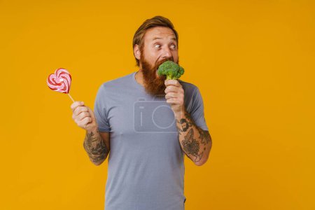 Foto de Hombre barbudo sosteniendo piruleta y brócoli mientras está de pie aislado sobre fondo amarillo - Imagen libre de derechos