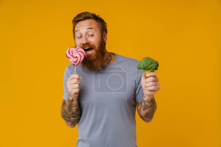 Photo for Bearded man holding lollipop and broccoli while standing isolated over yellow background - Royalty Free Image