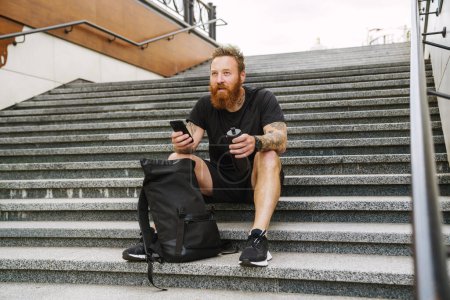 Photo for Young handsome bearded tattooed redhead man with bottle and phone in his hands looking aside, while sitting with backpack on the stairs outdoors - Royalty Free Image