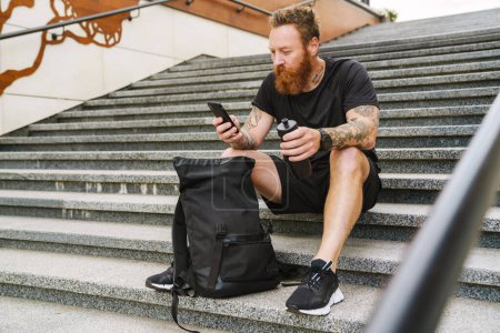 Téléchargez les photos : Jeune beau barbu tatoué rousse homme avec bouteille à la main tenant et utilisant son téléphone, tout en étant assis avec sac à dos sur les escaliers à l'extérieur - en image libre de droit