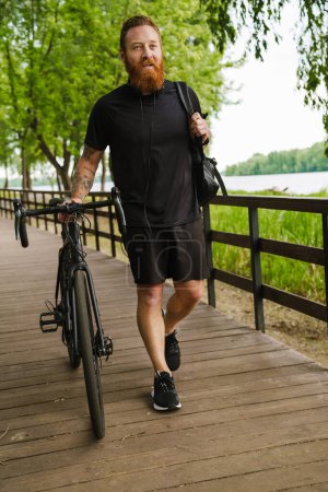 Photo for Adult handsome tattooed bearded calm man in black sport suit looking aside, while walking with bicycle on wooden bridge in park - Royalty Free Image