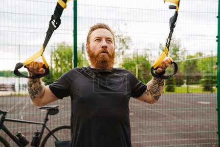 Foto de Joven guapo tatuado barbudo pelirrojo serio mirando hacia arriba y haciendo ejercicios con TRX cerca de la bicicleta en el campo de deportes al aire libre - Imagen libre de derechos