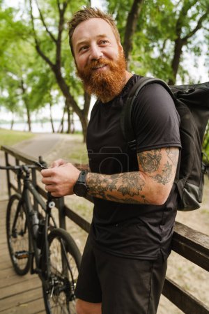 Photo for Portrait of young handsome smiling tattooed bearded man holding phone and looking aside, while leaning on wooden railing standing near bicycle in sunny park - Royalty Free Image