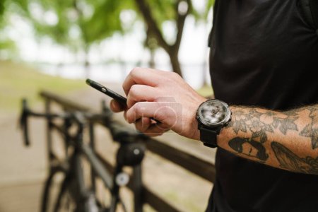 Foto de Macho tatuado mano con reloj sosteniendo el teléfono en el parque cerca de la bicicleta recortado, de cerca - Imagen libre de derechos