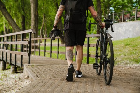 Téléchargez les photos : Portrait recadré à l'arrière d'un homme tatoué en costume de sport noir marchant à vélo dans un parc vert - en image libre de droit