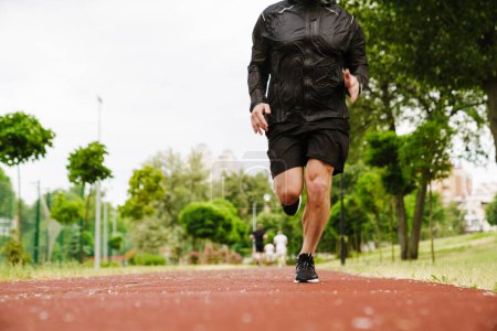 Téléchargez les photos : Portrait recadré de l'homme adulte en costume de sport noir qui court sur la piste à l'extérieur - en image libre de droit