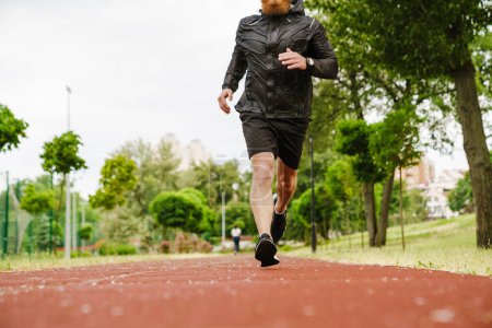 Foto de Retrato recortado de hombre pelirrojo barbudo adulto en traje deportivo negro corriendo en pista al aire libre - Imagen libre de derechos