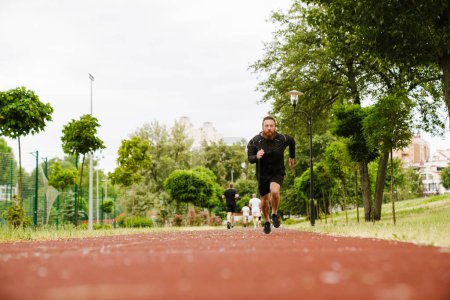 Photo for Portrait of adult bearded redhead man in black sport suit running on track outdoors - Royalty Free Image