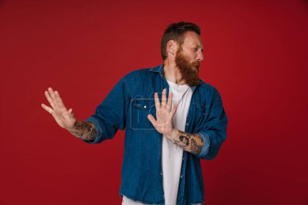 Photo for Adult bearded tattooed handsome redhead man doing rejection gesture with closed eyes and turning his head away, while standing over isolated red background - Royalty Free Image