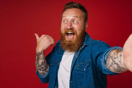 Photo for Selfie of adult bearded tattooed handsome smiling redhead man pointing and looking aside ,while standing over isolated red background - Royalty Free Image