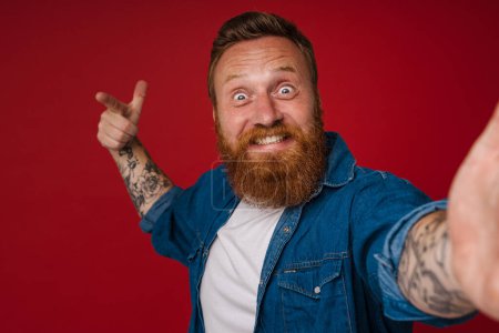 Foto de Selfie de adulto barbudo tatuado guapo entusiasta pelirrojo haciendo gesto de pistola con la mano levantada y los dedos mirando a la cámara, mientras está de pie sobre el fondo rojo aislado - Imagen libre de derechos