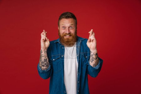 Photo for Adult bearded tattooed handsome hopeful redhead man with opened mouth doing hope gesture with crossed fingers and looking at camera , while standing over isolated red background - Royalty Free Image
