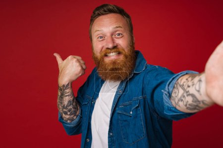 Téléchargez les photos : Selfie d'adulte barbu tatoué beau rousse souriant homme pointant de côté et regardant la caméra, tout en se tenant sur fond rouge isolé - en image libre de droit